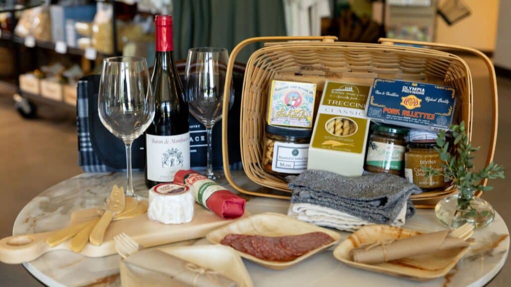 A picnic basket with wine, cheese, crackers, preserves, napkins, wine glasses, and wooden cutlery arranged on a round table indoors.