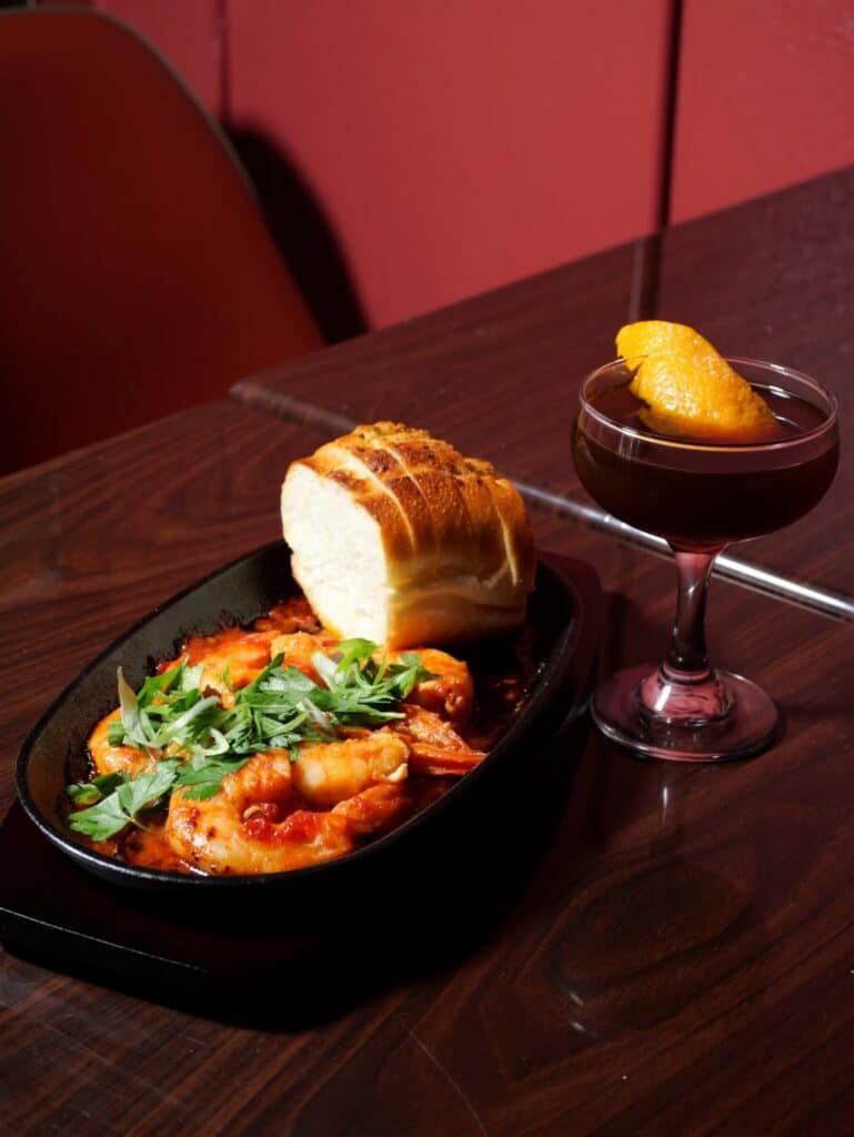 A plate of shrimp in tomato sauce with fresh herbs and a slice of bread, served next to a cocktail garnished with an orange peel on a dark wooden table.