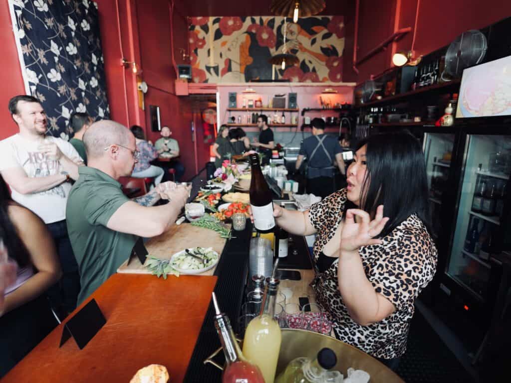 A bartender hands a bottle to a customer at a busy, colorful bar with patrons seated, drinks and ingredients on the counter, and staff working in the background.