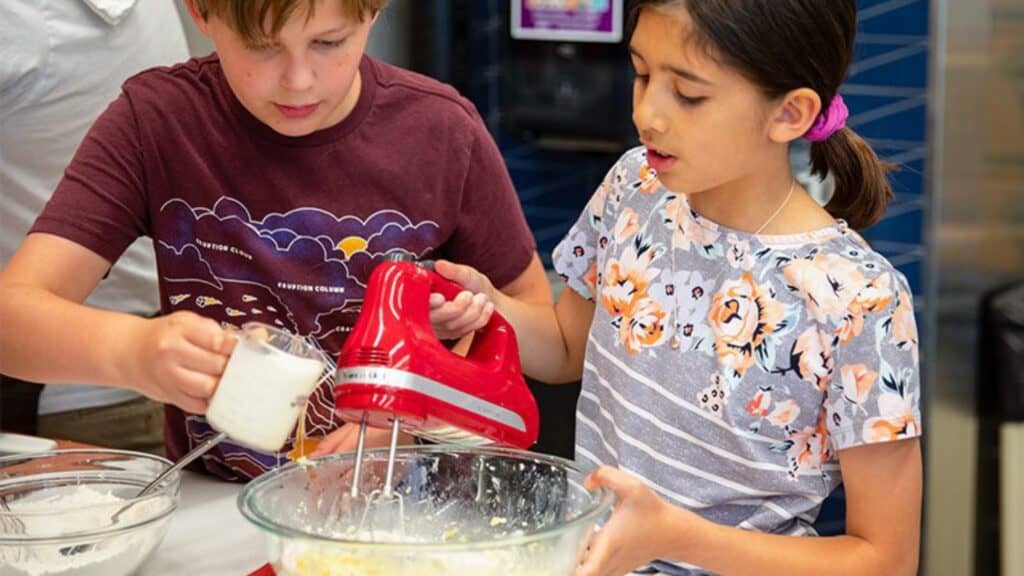 Two children make batter; one pours milk while the other uses a red electric hand mixer in a glass bowl, with baking ingredients on the table.