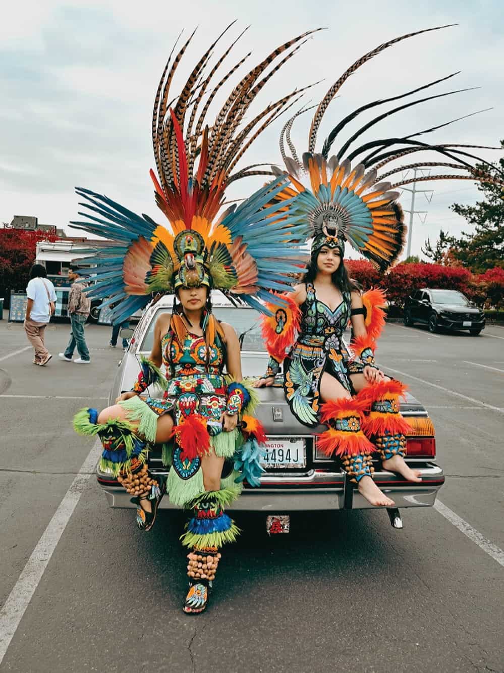 Two people in colorful, elaborate feathered costumes sit on the trunk of a car in a parking lot, capturing a vivid moment of Mexican culture in Seattle. Trees and buildings are visible in the background, adding context to this unique scene that could easily be part of a Seattle photo essay.