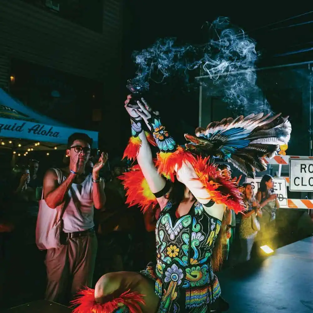 Performer in colorful feathered costume lifts a smoking vessel during a nighttime outdoor event, celebrating Mexican culture in Seattle, with spectators and road closure signs in the background.