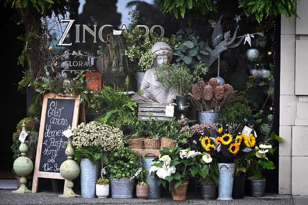 Flower shop entrance with various potted plants, sunflowers, a chalkboard sign, and a decorative statue displayed outside—evoking the charm of a European-inspired getaway just steps away from Seattle.
