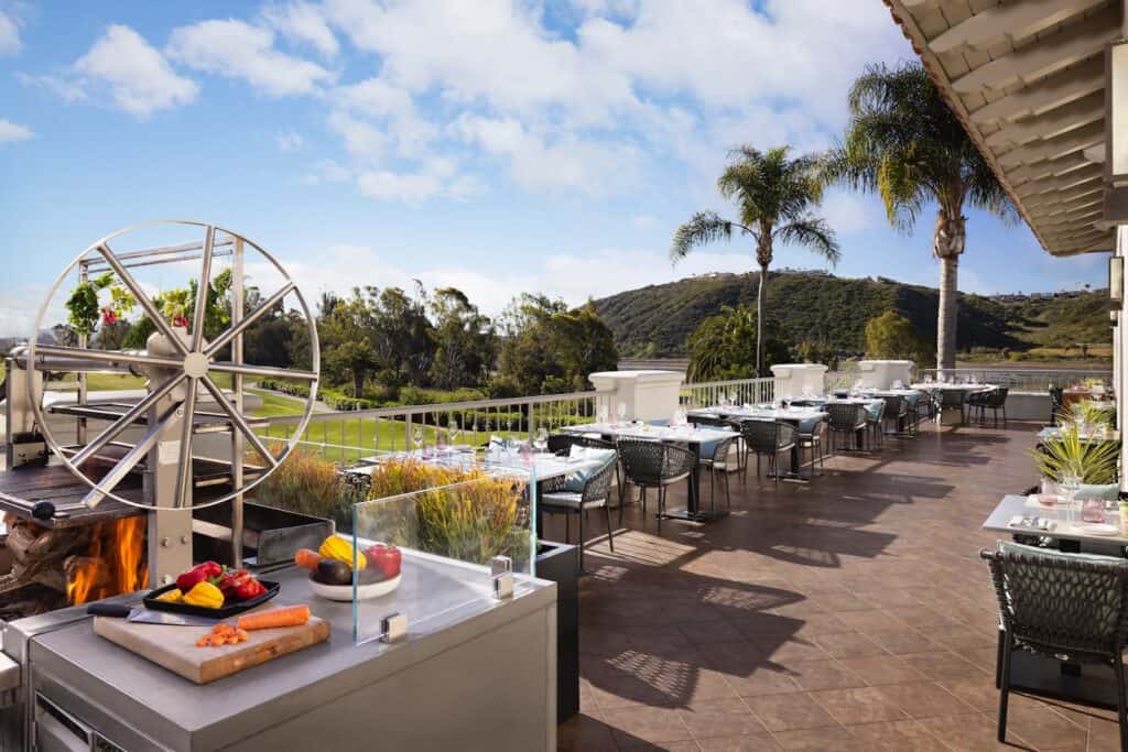 Outdoor restaurant terrace with tables set for dining, a modern grill station in the foreground, and a scenic view of green hills and palm trees under a partly cloudy sky.