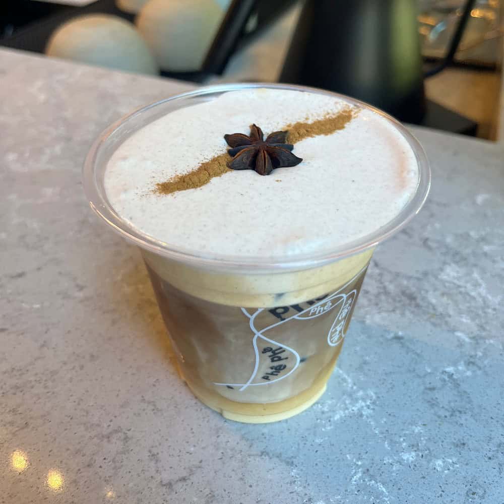A plastic cup of iced coffee with a thick layer of foam, topped with a line of cinnamon and a star anise, sits on a light gray countertop.