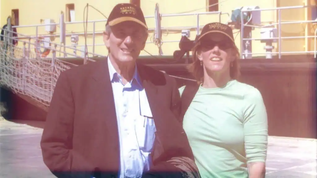 Two people wearing dark caps stand together in front of a yellow boat or ship, posing for a photo outdoors on a sunny day.