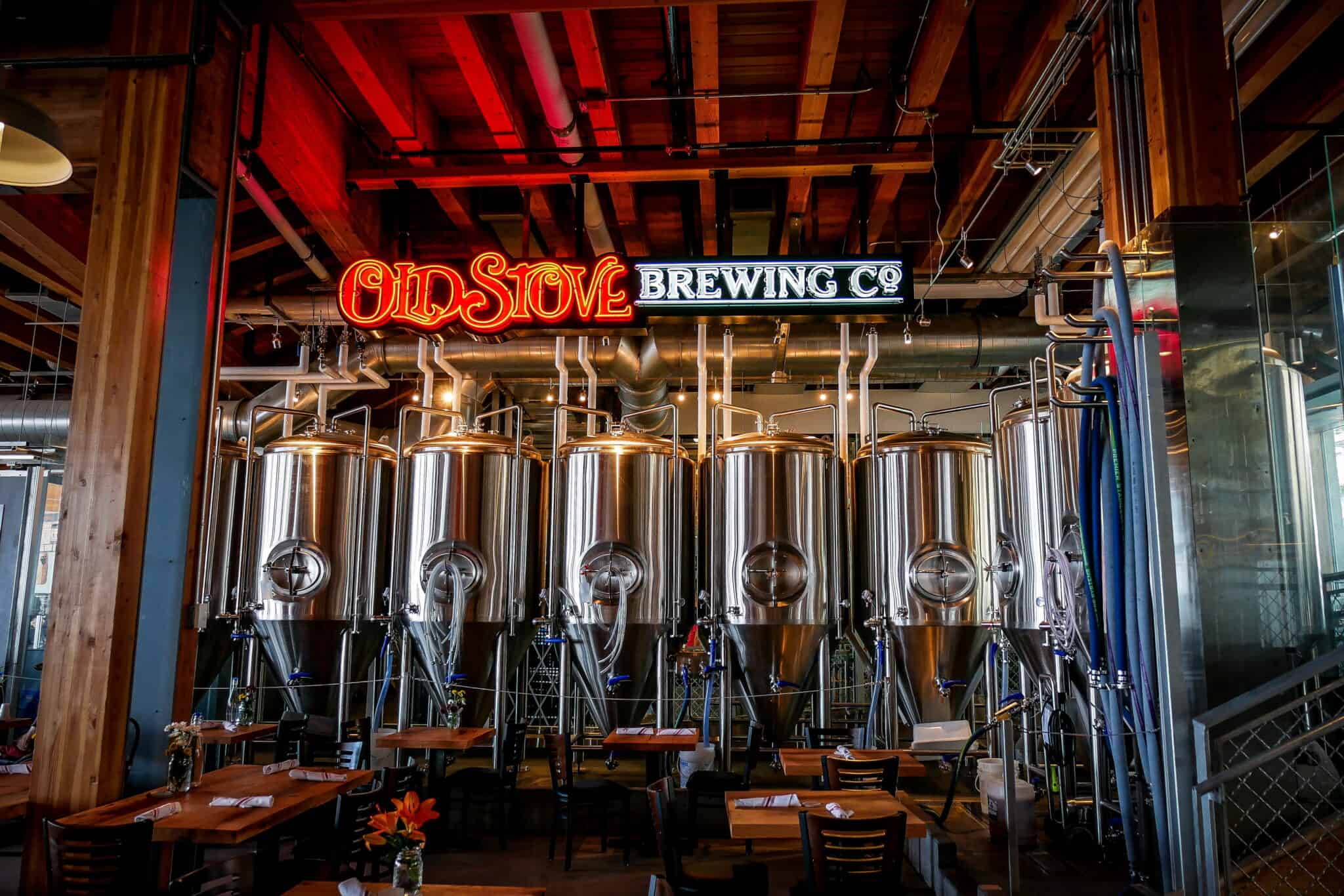Six large stainless steel brewery tanks stand behind tables and chairs beneath a neon "Old Stove Brewing Co" sign, offering guests a warm welcome and cold beer in an inviting industrial-style brewery.