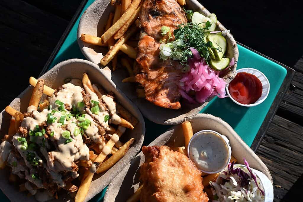 Three takeout trays with fries: one topped with grilled chicken and pickled vegetables, one with cheese sauce and green onions, and one with fried fish, coleslaw, and dipping sauces.