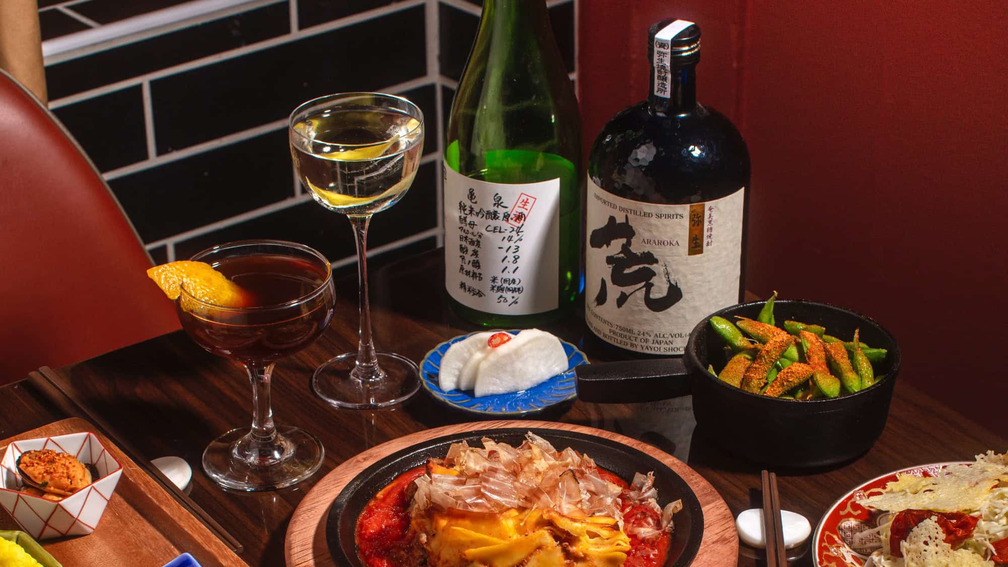A table set with various Japanese dishes, including okonomiyaki, edamame, pickled radish, cocktails, and bottles of sake and liquor against a black tiled and red wall background.