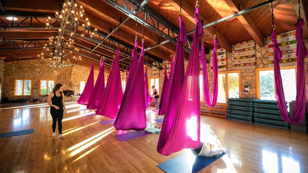 A spacious studio with wooden floors and ceiling features purple aerial yoga hammocks, yoga mats, stacked blocks, and two people preparing for a class. Sunlight streams through large windows.