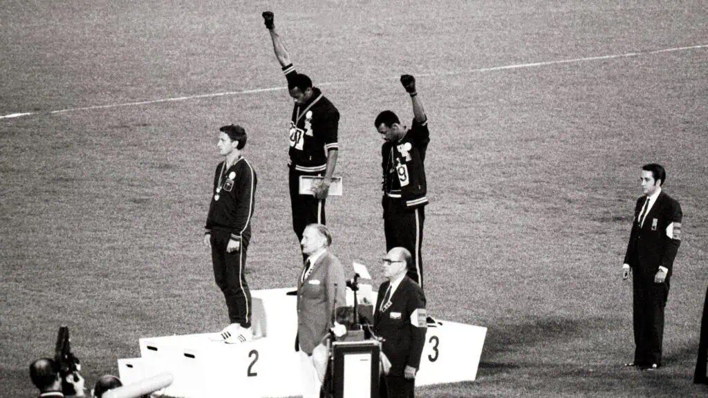 Three athletes stand on an Olympic podium; two raise their fists while the third stands with his hands down. Several officials and photographers are present.