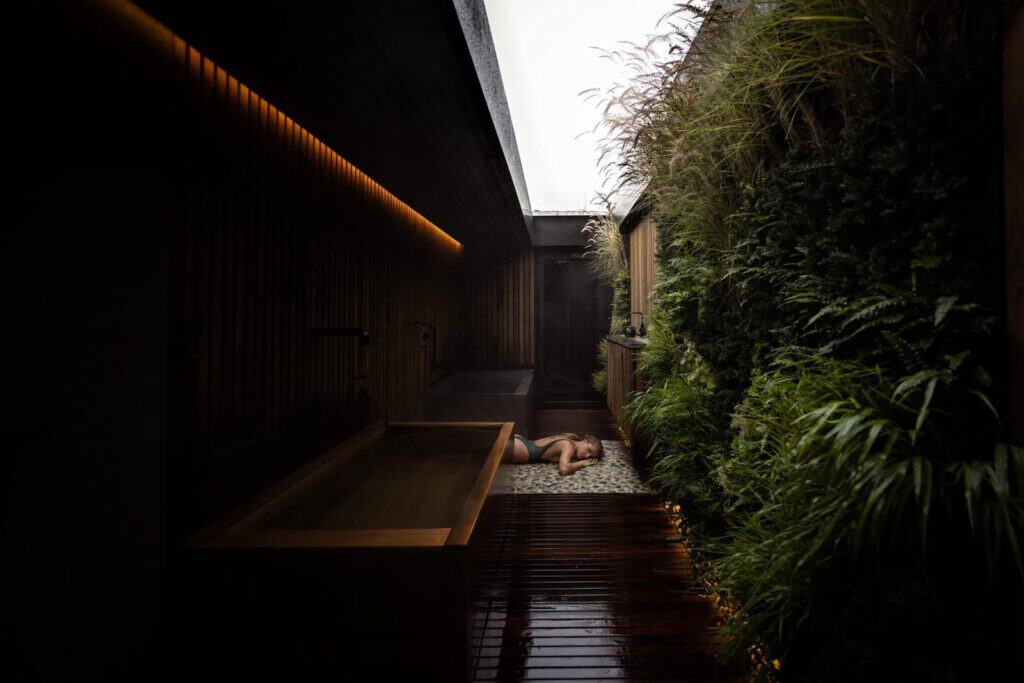 A person lies on a stone floor in a narrow outdoor bath area with wooden walls and lush green plants along one side.