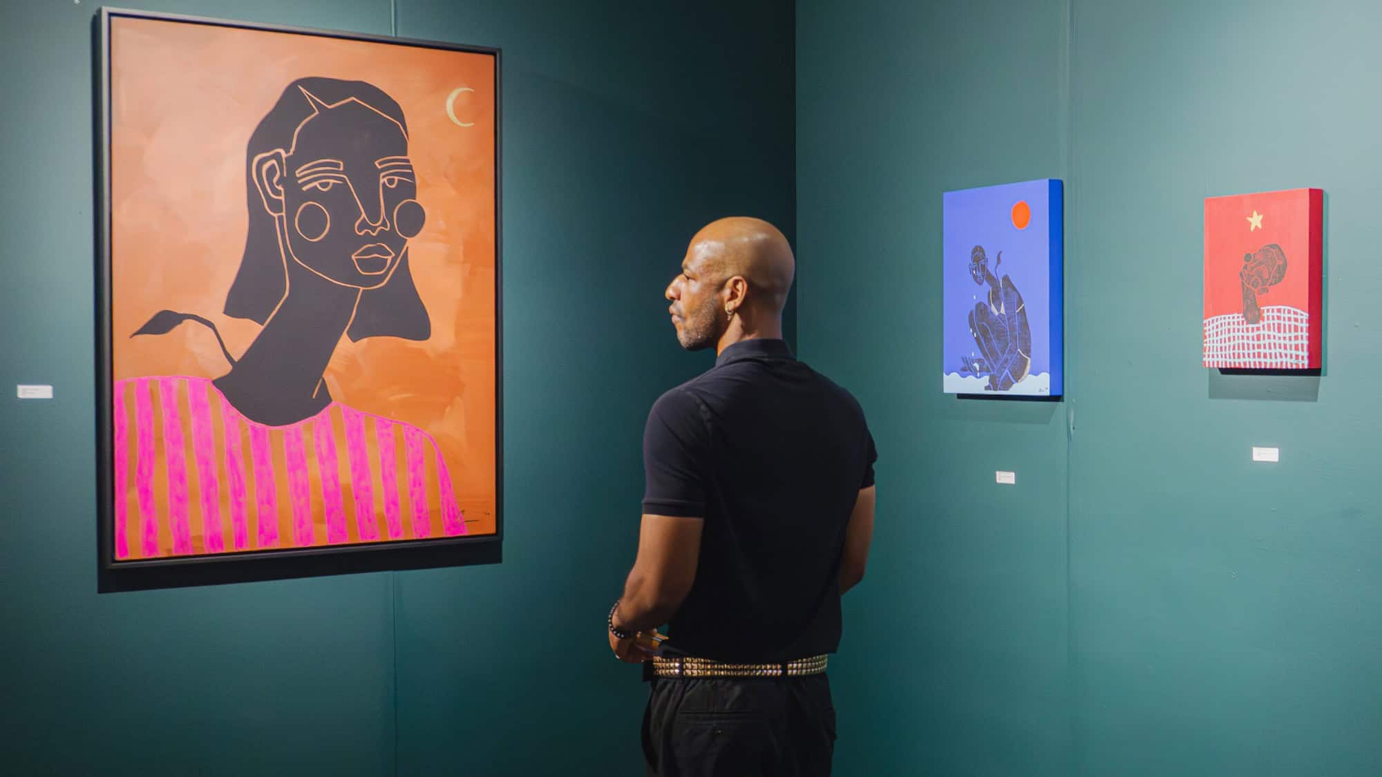 A person stands in an art gallery, viewing colorful paintings of abstract human figures displayed on a green wall.