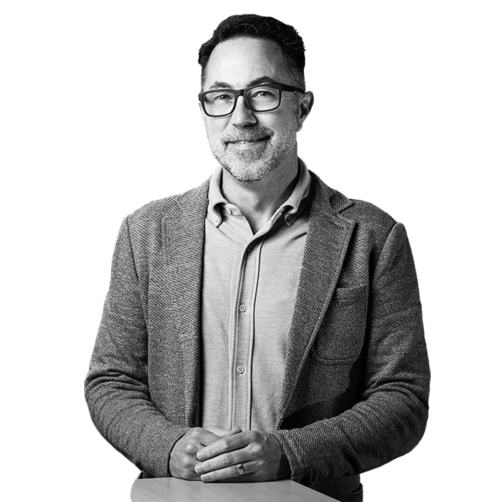 A man with glasses, a beard, and short hair, wearing a buttoned shirt and textured blazer, stands with hands resting on a wooden surface against a plain white background—evoking the thoughtful precision found in Pacific Northwest medical facility design.