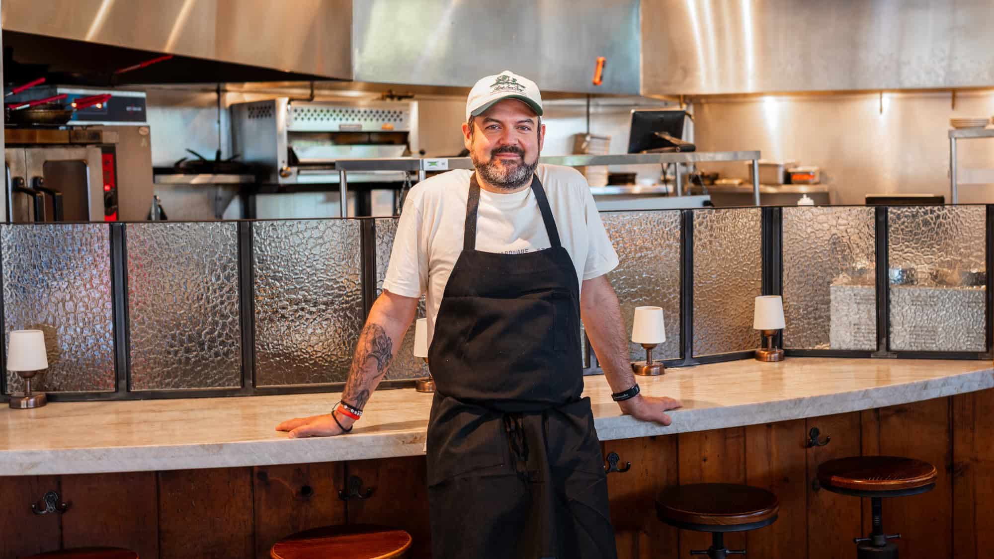 A man wearing a white cap, t-shirt, and black apron stands behind a counter in a commercial kitchen, facing the camera and smiling.