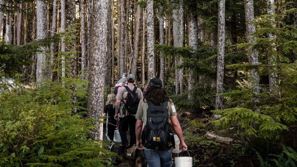 Four people carrying buckets and backpacks walk in single file along a narrow trail through a dense, green forest filled with tall trees.