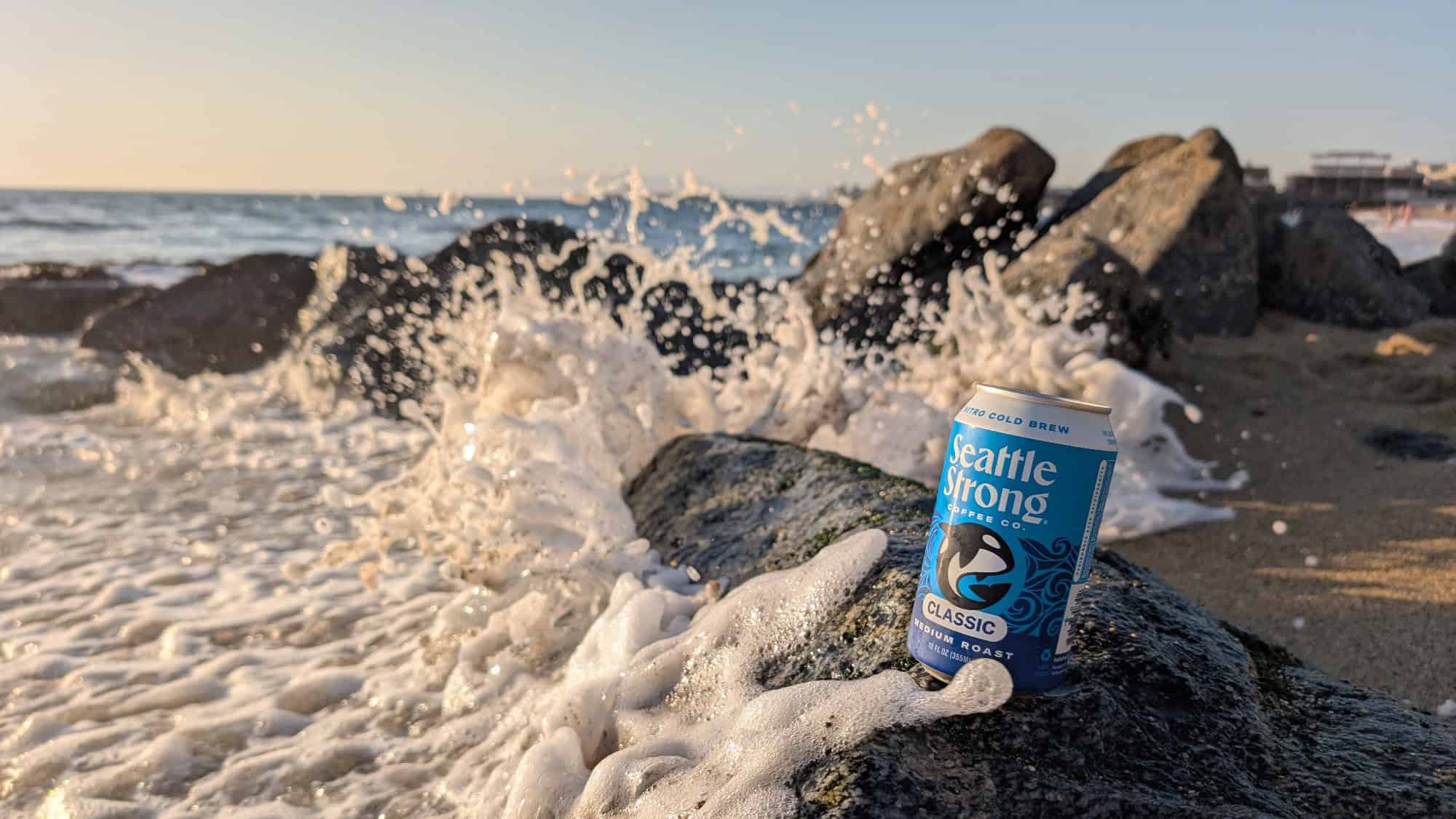 A Seattle Strong cold brew can sits on a rocky beach as ocean waves crash and splash nearby under a clear sky.