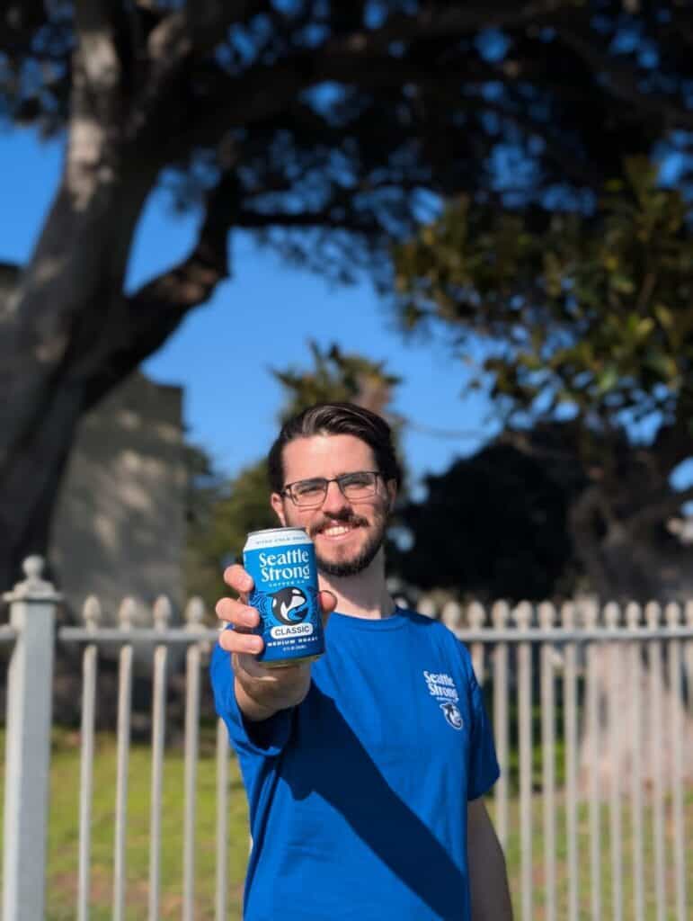 Man in a blue Seattle Strong Coffee shirt holds a can of Seattle Strong Classic Cold Brew forward, standing outdoors by a metal fence with trees in the background.