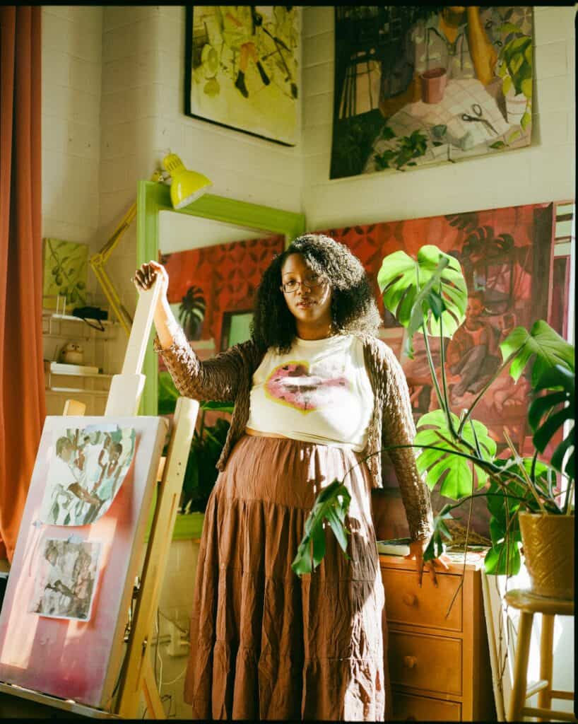 A woman stands in an art studio surrounded by plants, paintings, and natural light, with one hand resting on an easel displaying artwork.