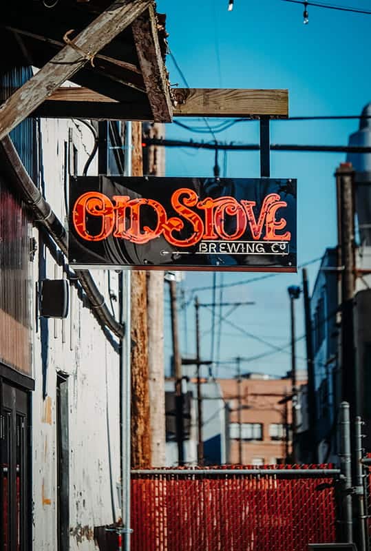 A neon sign reading "Old Stove Brewing Co" offers a warm welcome above an alley, with industrial buildings in the background promising cold beer within.