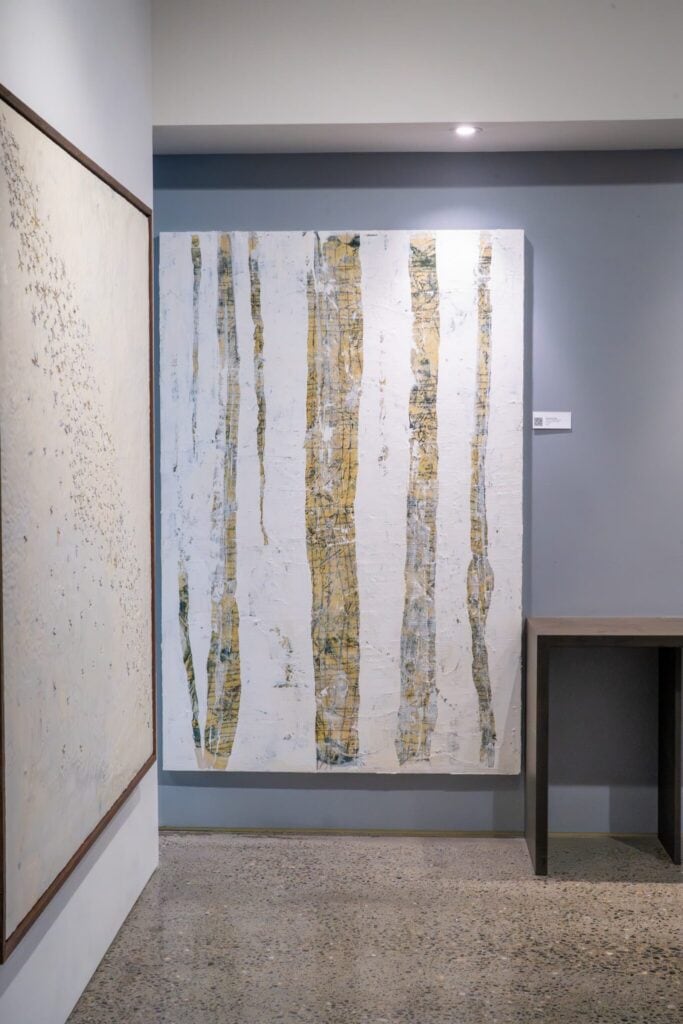 Large abstract painting with vertical yellow and white streaks hangs on a gray wall near a wooden table and another artwork in a modern gallery space.