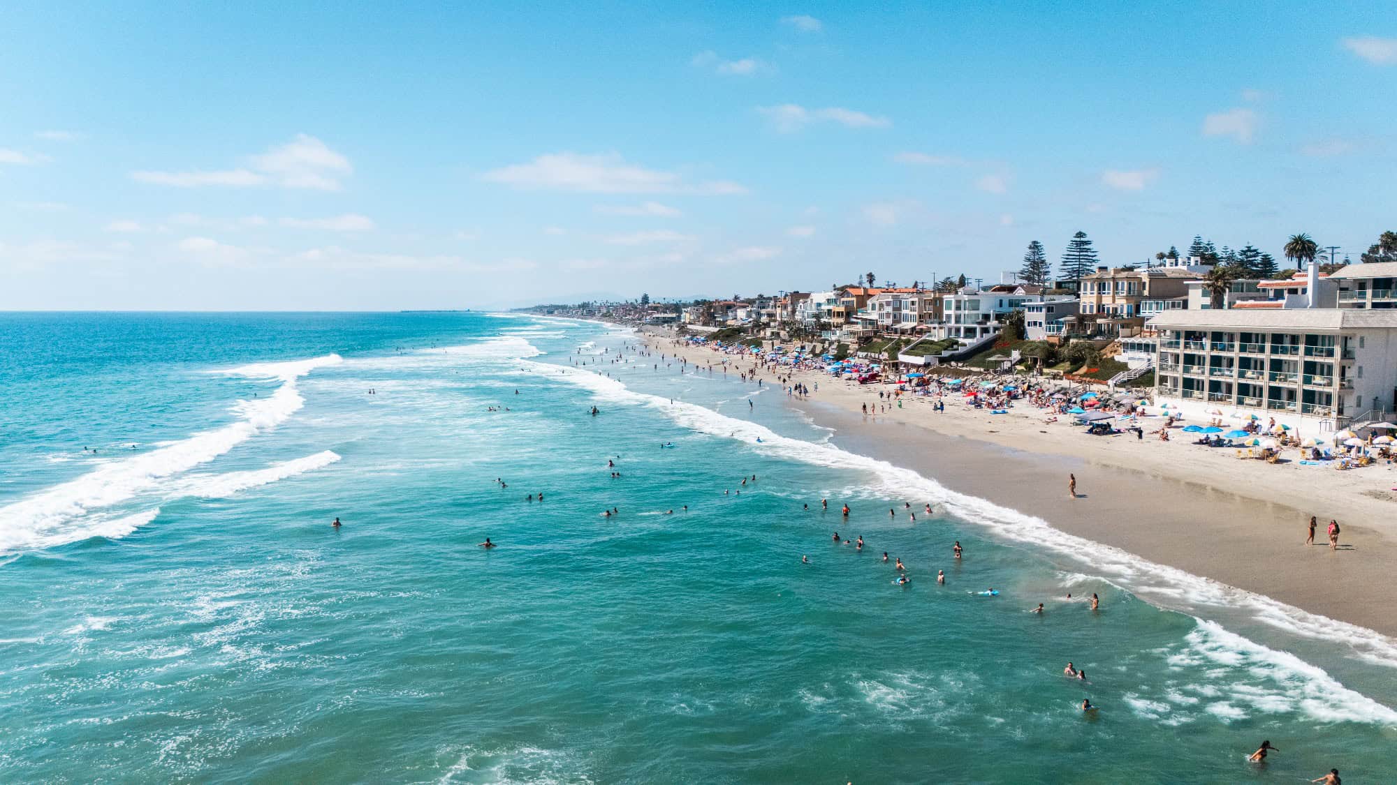 A wide sandy beach with people swimming in the ocean and sunbathing near buildings and houses along the shoreline under a blue sky.