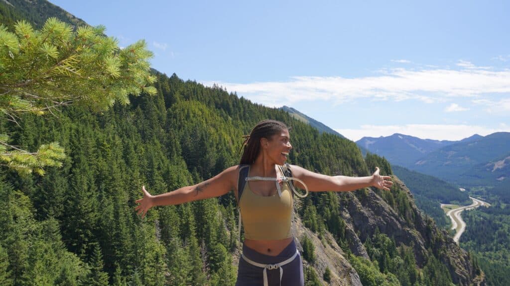 Person standing with arms outstretched on a mountain, surrounded by green forest and distant peaks under a clear blue sky.