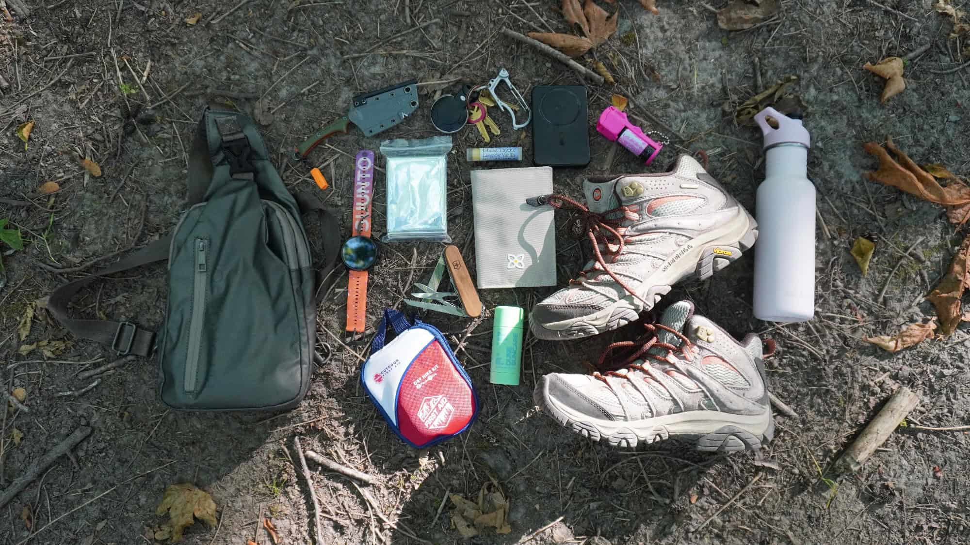 Hiking gear on ground: backpack, first aid kit, knife, compass, flashlight, water bottle, hiking boots, whistle, sunscreen, bug spray, map, lighter, gloves, watch, and small tools.