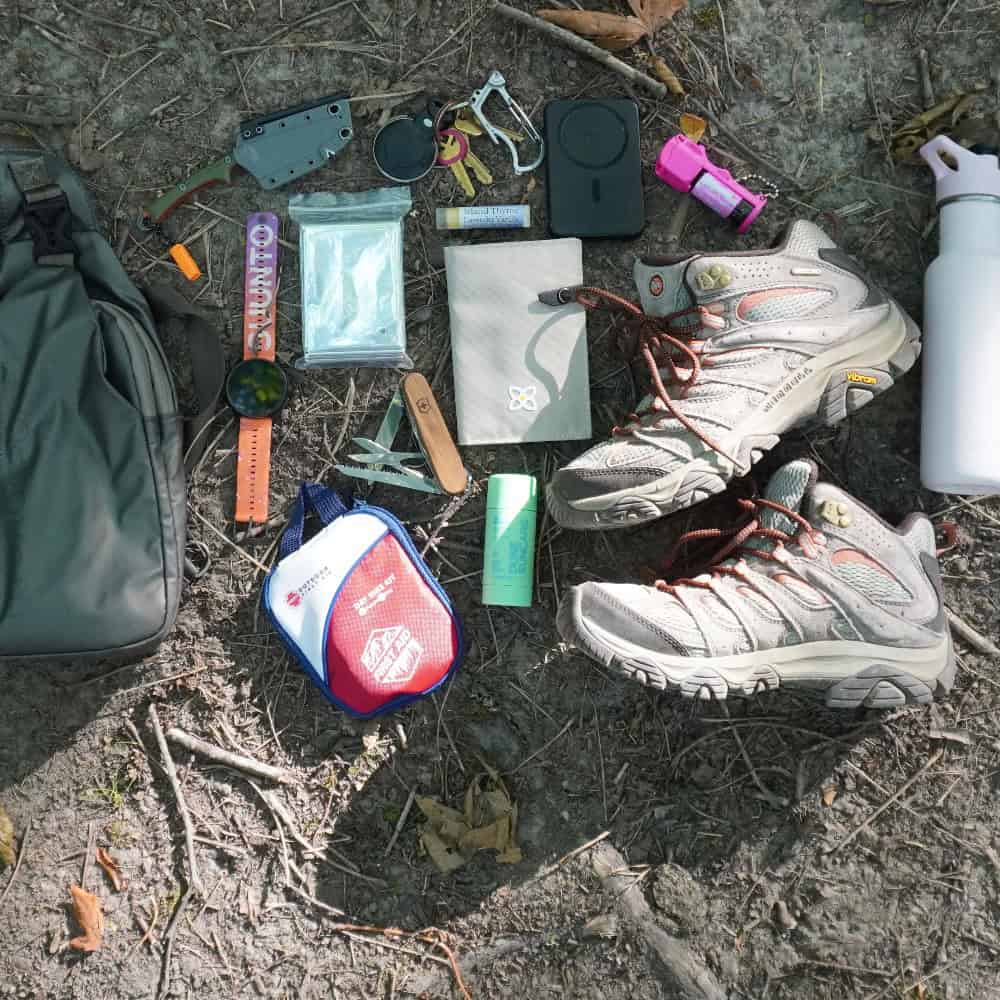 Hiking essentials laid out on the ground, including shoes, water bottle, bag, first aid kit, sunscreen, map, compass, tools, flashlight, and other small gear.