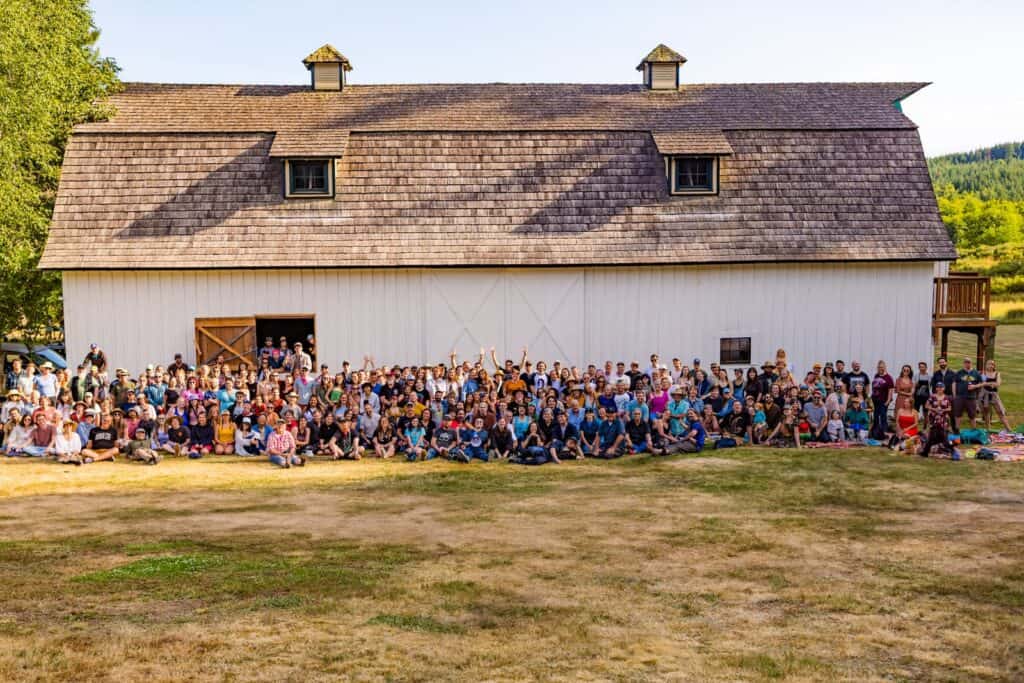A large group of people poses for a photo in front of a white barn on a sunny day, with trees and hills in the background.
