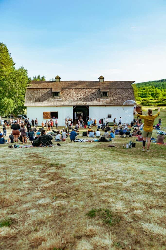 People gather on a grassy hill in front of a large white barn for an outdoor event on a sunny day, with someone playing frisbee in the foreground.
