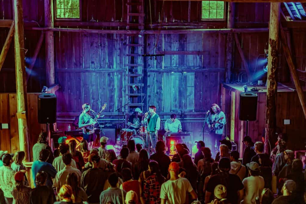 A band performs on stage inside a wooden barn while a crowd of people watches. Stage lights cast colorful glows throughout the rustic venue.