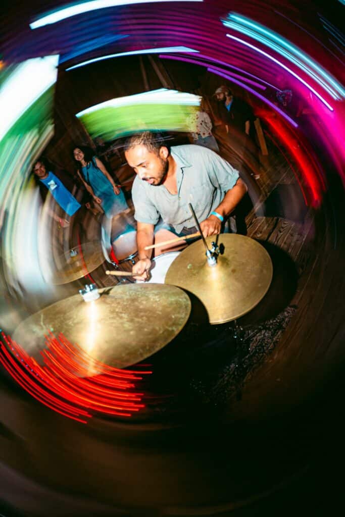 A man plays drums energetically onstage, with colorful light trails swirling around him and people watching in the background.
