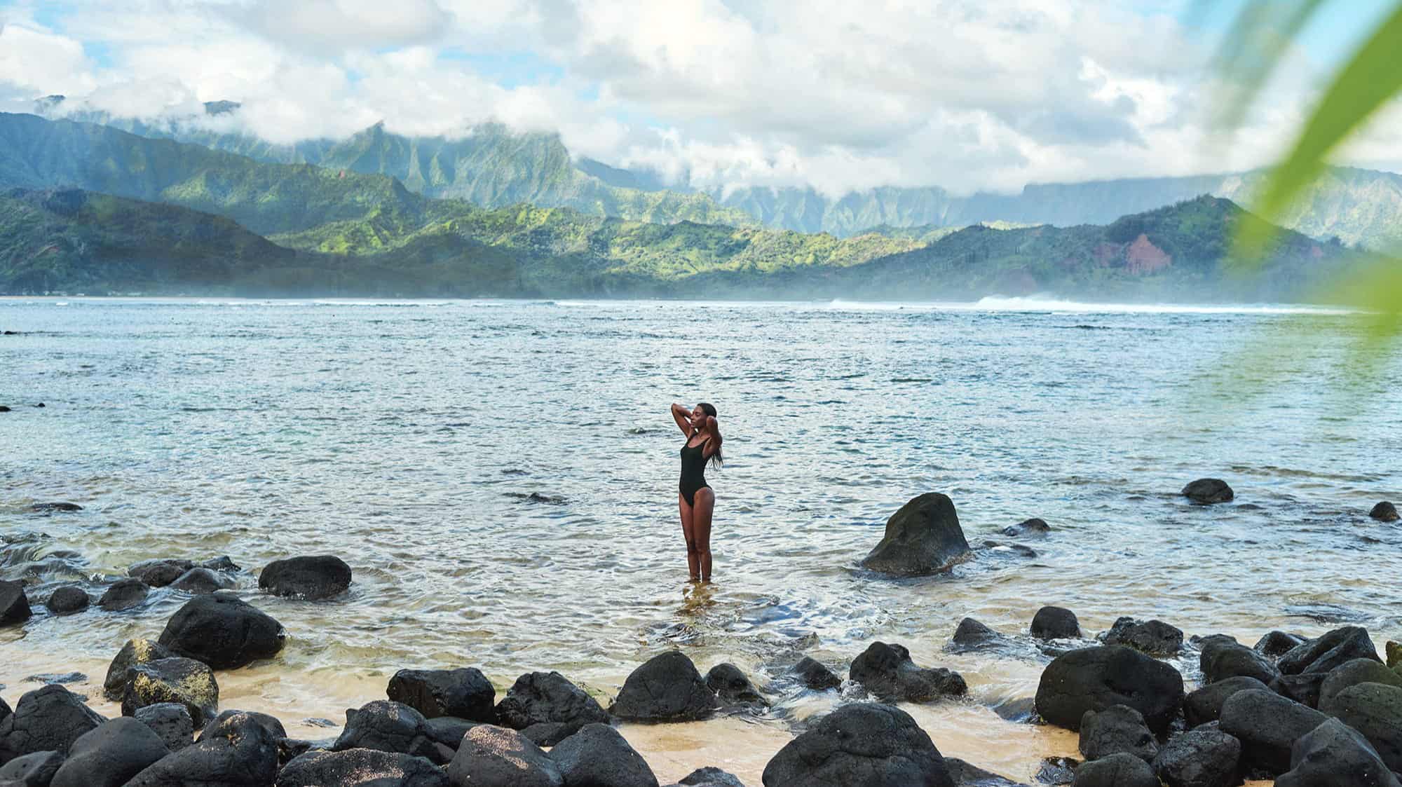 A person in a black swimsuit stands among rocks in shallow water, with mountains and a cloudy sky in the background—evoking the tranquil vibe of wellness retreats at 1 Hotel Hanalei Bay.