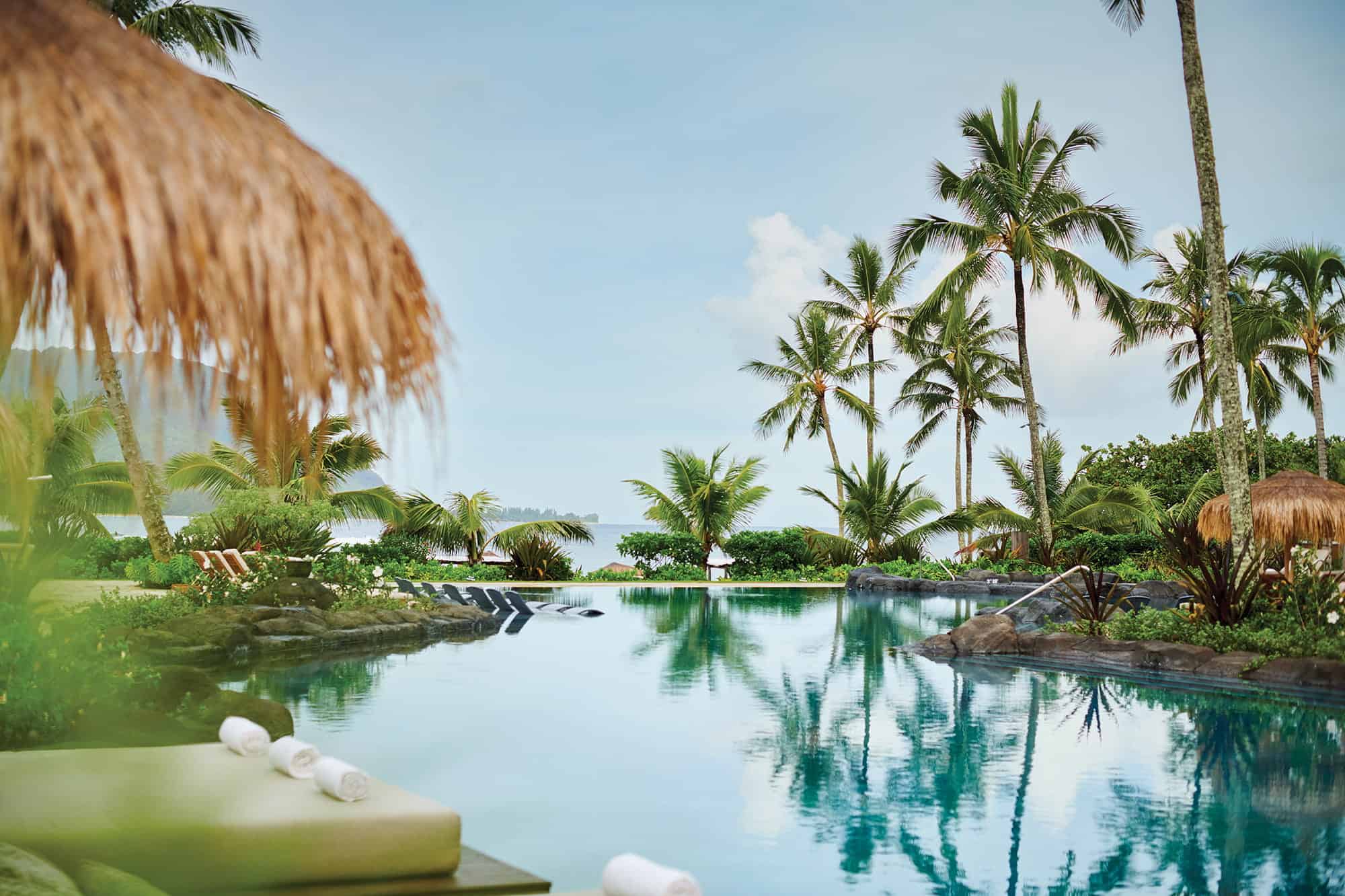 Resort swimming pool surrounded by palm trees, lounge chairs, and thatched umbrellas at 1 Hotel Hanalei Bay in Kauai, offering ocean views and a serene setting ideal for wellness retreats under a clear sky.