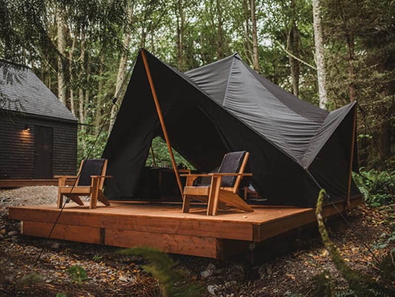 A black canvas tent with wooden supports sits on a wooden platform in a forest, with two wood chairs placed in front—an inviting scene that embodies the tranquil atmosphere of an Olympic Peninsula wellness retreat, just a short drive from destinations like Sol Duc Hot Springs and Soak on the Sound Port Townsend.