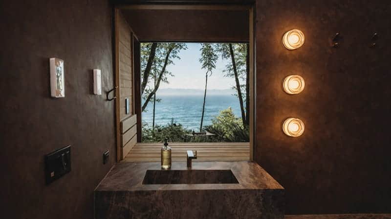Modern bathroom with a stone sink, soap dispenser, and lit wall lights, featuring a large open window with a view of trees and the ocean—perfect for those seeking an Olympic Peninsula wellness retreat.