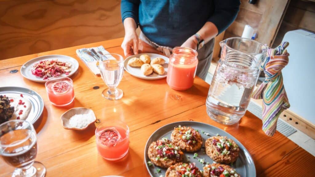 A person stands at a wooden table set for a cozy breakfast, complete with plates of food, pink juice, a lit candle, and water—capturing the relaxed vibe of craft camp for grown-ups who love adult crafts and good company.