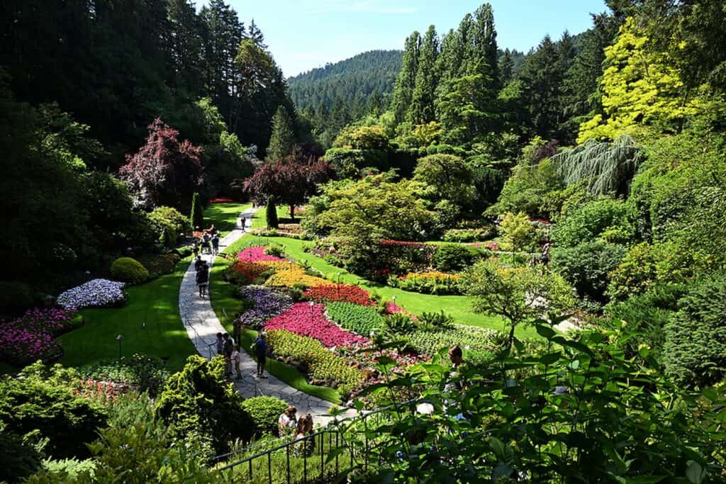 A landscaped garden with winding paths, colorful flowerbeds, green lawns, and people walking among trees and shrubs on a sunny day. Forested hills appear in the background.