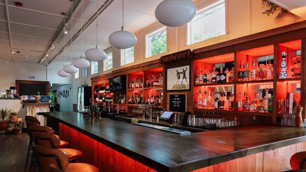 A modern bar with a wooden counter, shelves stocked with various liquor bottles, bar stools, and pendant lights hanging from the ceiling.