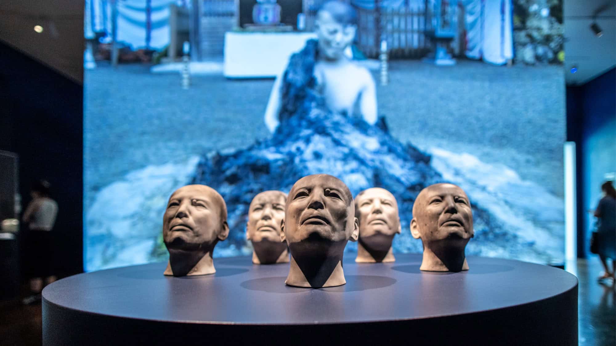 Five sculpted human heads are displayed on a round pedestal with a blurred video projection of a person and outdoor scene in the background.
