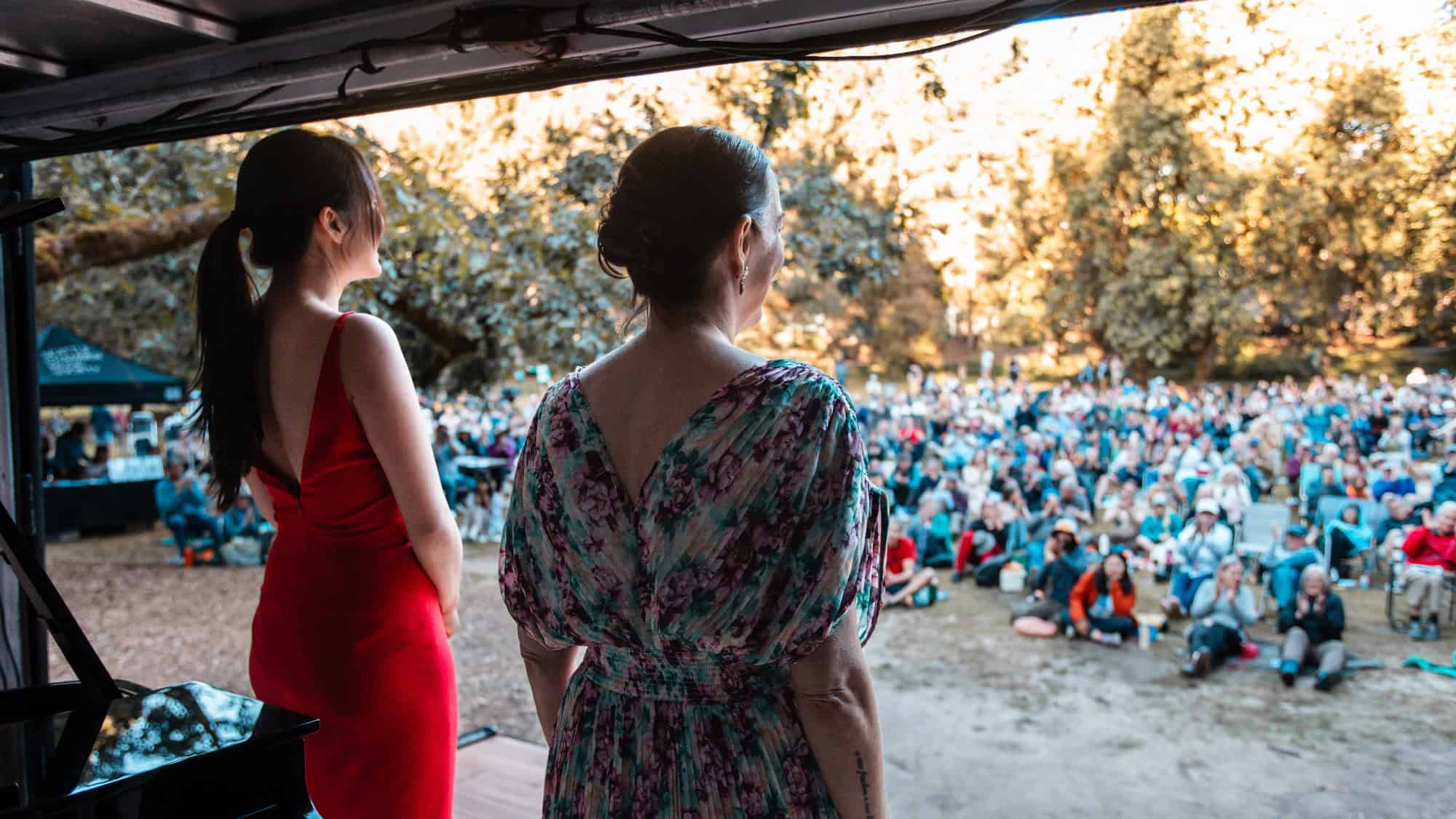 Two women stand on an outdoor stage facing a large seated audience in a park during the day.