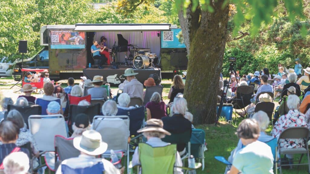 A crowd seated on lawn chairs watches a live outdoor music performance on a small stage in a park, surrounded by trees on a sunny day.