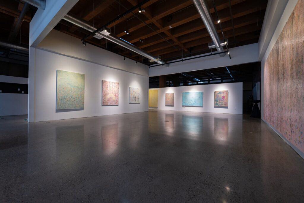 A contemporary art gallery with polished concrete floors, white walls, and various large abstract paintings displayed under track lighting.
