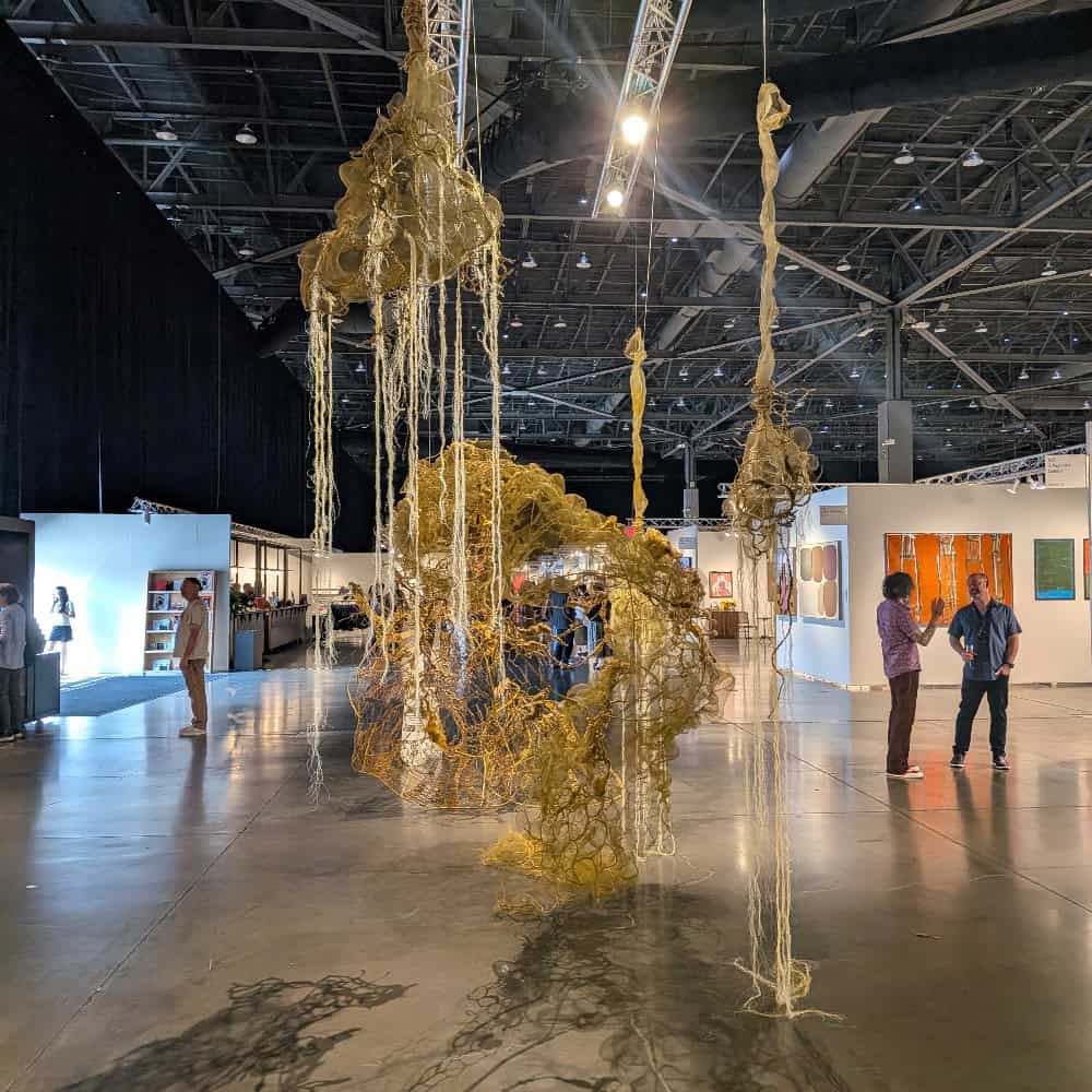 Large abstract sculpture made of tangled fibers hangs from the ceiling in a spacious art gallery. Several people observe the artwork and paintings displayed on the walls.