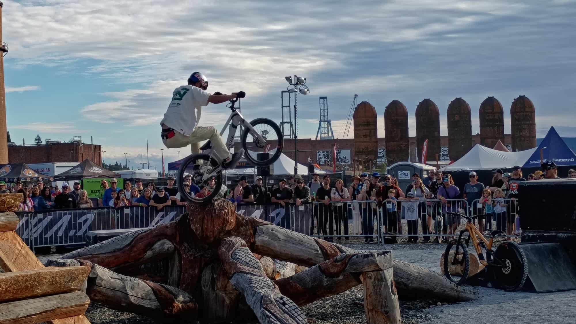 A BMX rider jumps over stacked logs during an outdoor event, while a crowd watches behind metal barriers under a partly cloudy sky.