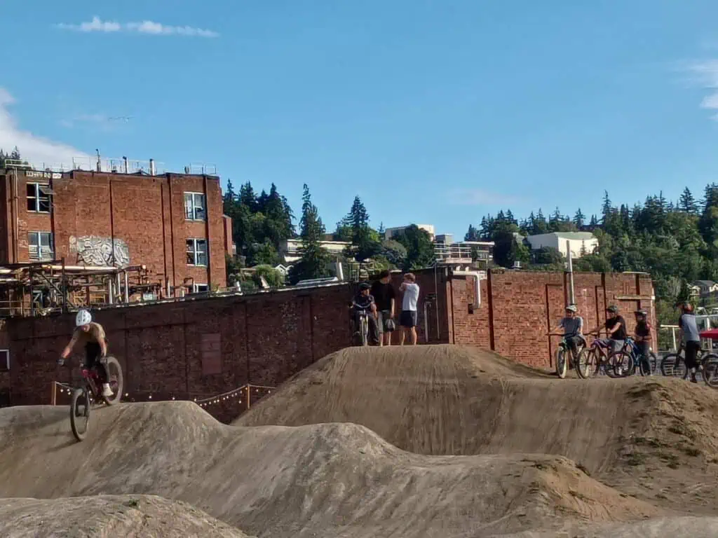 Several cyclists ride on a dirt pump track while others watch nearby; brick buildings and trees are visible in the background under a clear sky.