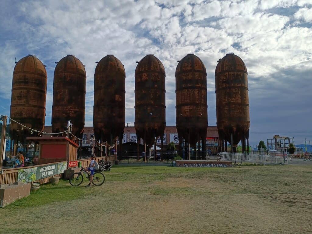 Six large, rusted silos stand in a row on grass with a small stage and wooden structures nearby under a partly cloudy sky.