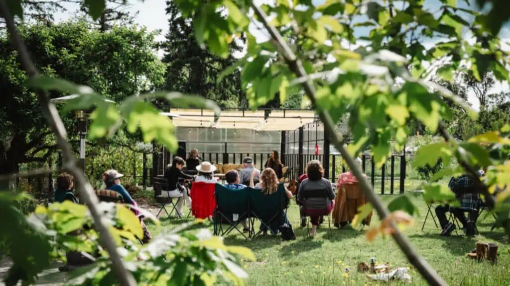 A group of people sit on lawn chairs in a circle outdoors near a glass building, surrounded by trees and greenery.