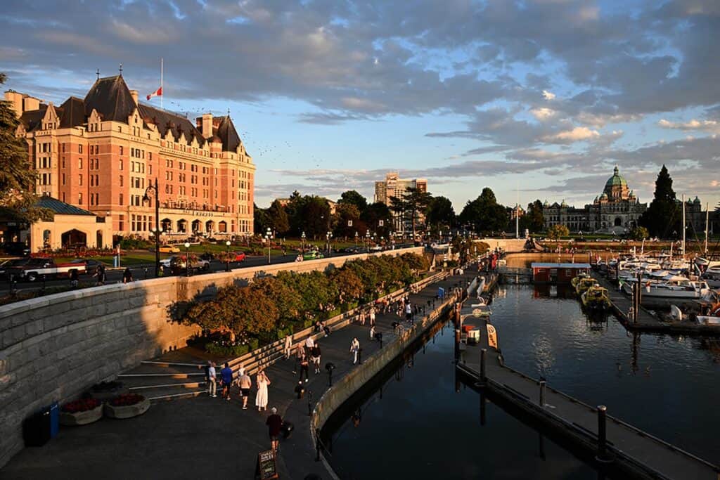 People walk along a waterfront promenade beside a historic hotel, with boats docked in the marina and a domed government building visible in the distance under a partly cloudy sky.